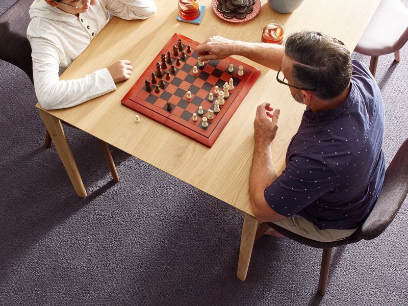 Two people playing chess at a wooden table in a room with purple carpet from Carpet & Flooring By Denny Lee in Abingdon, MD